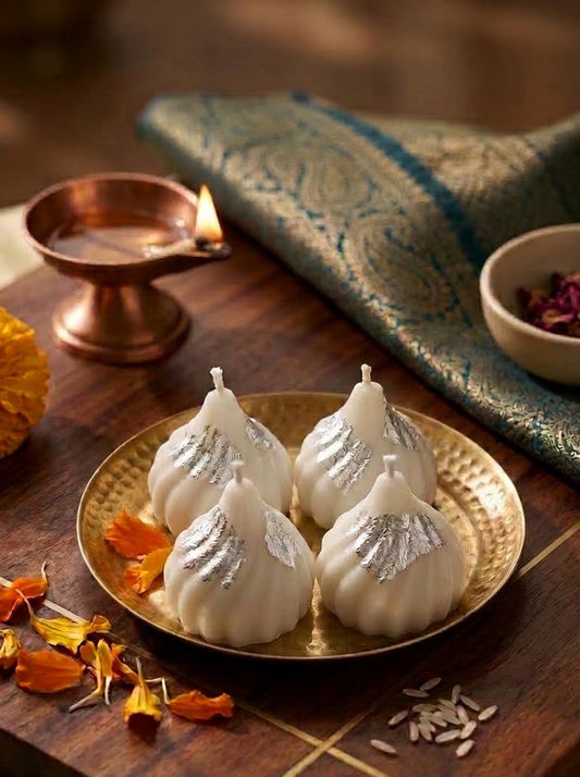 Four white wax candles shaped like sweets, decorated with silver foil leaves, arranged on a gold-colored plate. The plate is set on a wooden tray alongside a lit copper diya, a patterned blue and gold fabric napkin, and dried orange flower petals.