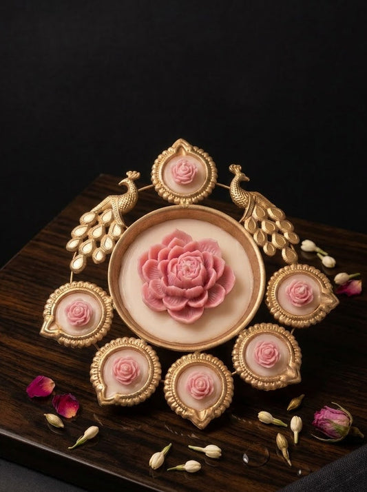 An ornate gold-toned decorative brooch or jewelry piece featuring a central pink rose surrounded by six smaller rose motifs and peacock-like feather details, displayed on a dark wooden surface with scattered rose petals and buds.