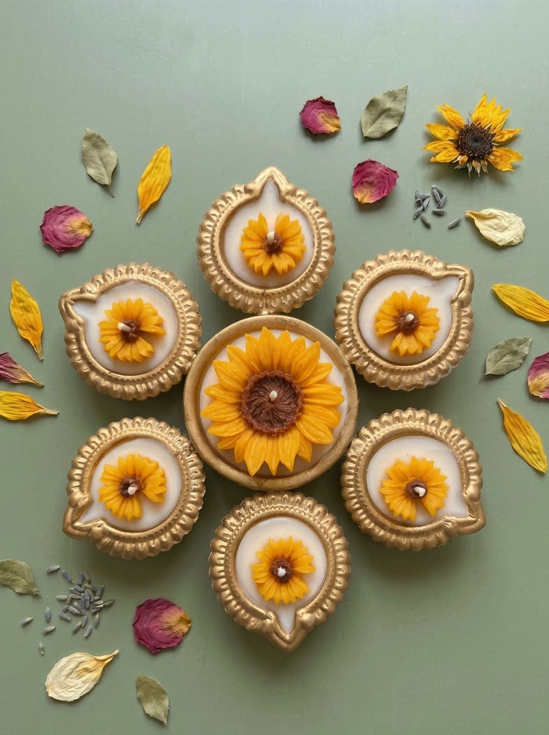 A decorative flat-lay arrangement of six gold-toned diya-style holders with yellow flower inlays, arranged in a circular pattern on a muted sage green background with scattered dried petals and leaves.