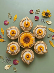 A decorative flat-lay arrangement of six gold-toned diya-style holders with yellow flower inlays, arranged in a circular pattern on a muted sage green background with scattered dried petals and leaves.