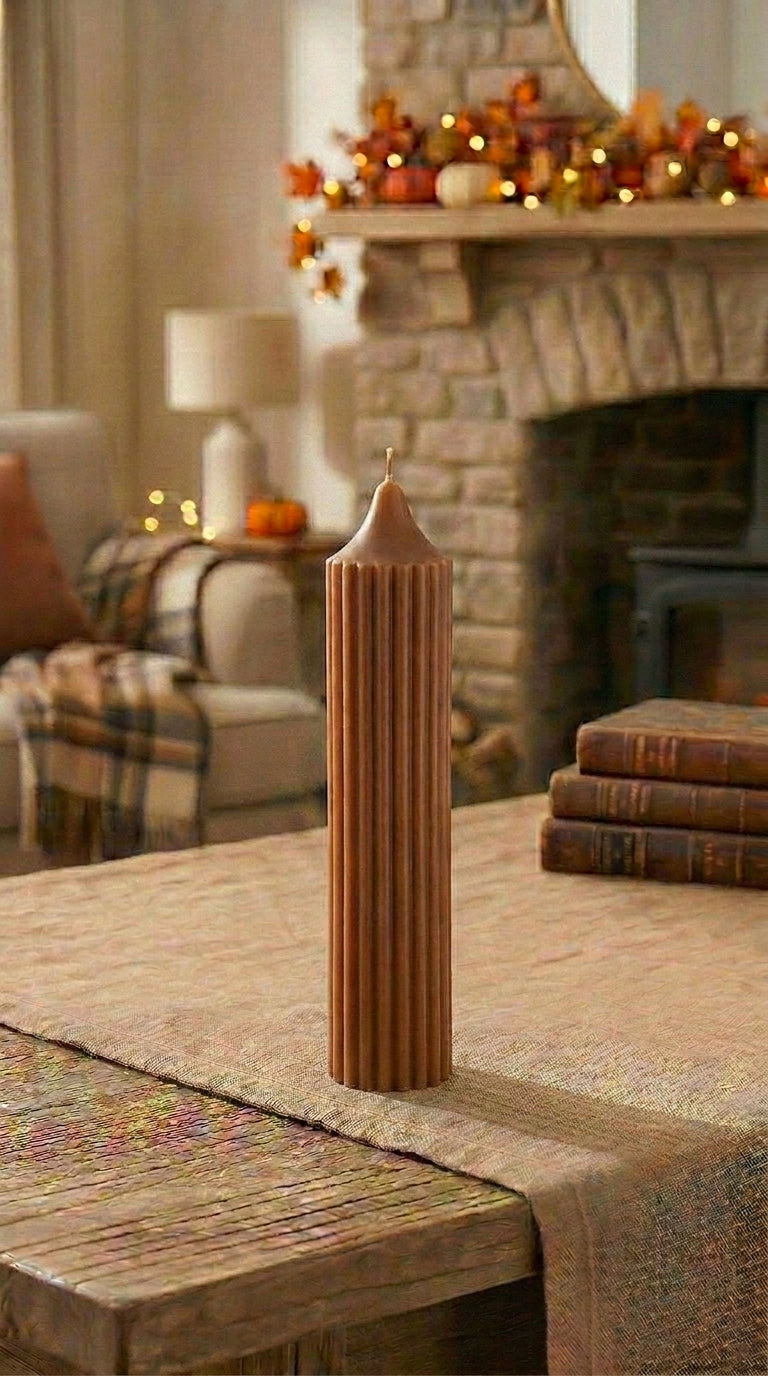 Tall brown candle on a wooden table with a stone fireplace and decorative pumpkins in the background.
