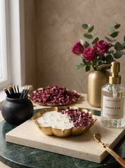 A clear glass bottle of Maison de Crève Embalance Oil with a gold pump, styled on a marble tray with dried rose petals, white candles, and a gold vase of roses.