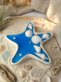 Decorative blue starfish with white shells on a sandy surface with seashells and driftwood.