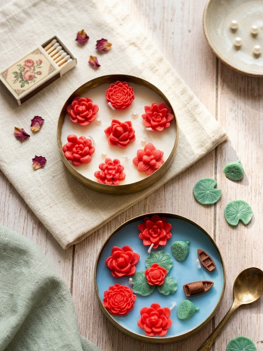 Two decorative plates with gold-toned rims, one cream and one light blue, each holding red flower-shaped candles and small green lily pads, arranged on a rustic wood surface with a matchbox, dried petals, a spoon, and a small dish.