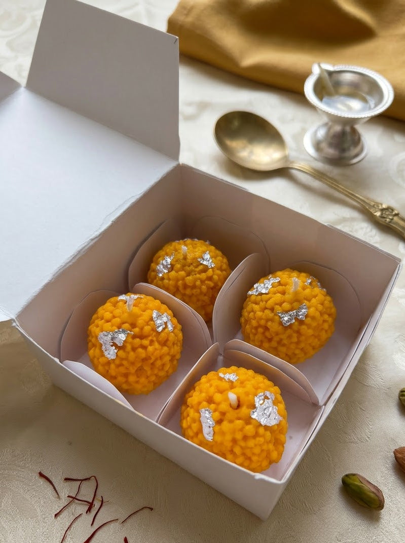 Four golden-yellow Indian sweet rice balls (laddoo) decorated with silver foil, presented in an open white cardboard box on a patterned cream tablecloth with a gold spoon, silver cup, and almonds nearby.