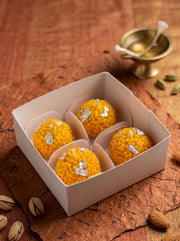 Four golden-yellow sweet rice balls (laddoo) with silver foil accents, presented in a white square box on a rustic brown textured surface with almonds and a small brass bowl in the background.