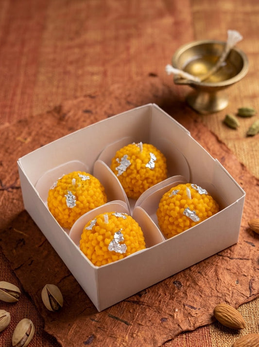 Four golden-yellow sweet rice balls (laddoo) with silver foil accents, presented in a white square box on a rustic brown textured surface with almonds and a small brass bowl in the background.