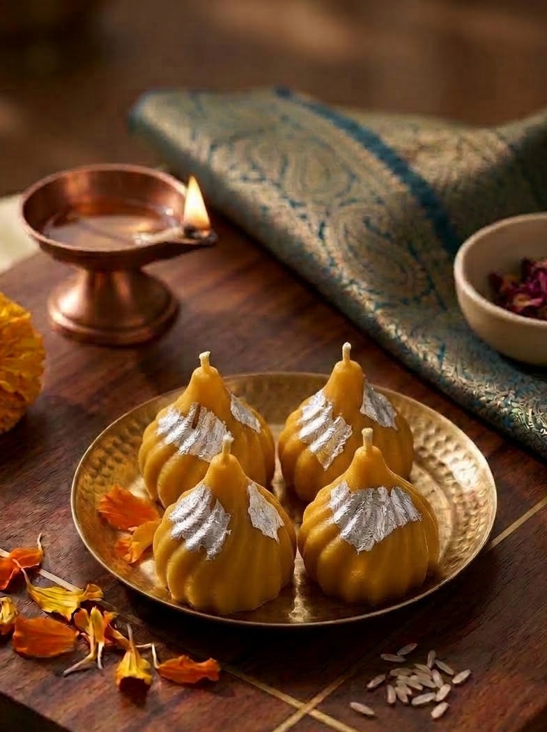 Four golden-yellow Indian sweets, likely ladoo or modak, decorated with silver powder, arranged on a brass plate on a wooden board. The setting includes a copper diya, a patterned green textile, and flower petals.