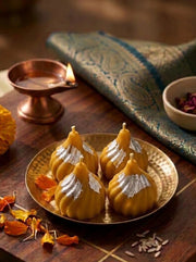 Four golden-yellow Indian sweets, likely ladoo or modak, decorated with silver powder, arranged on a brass plate on a wooden board. The setting includes a copper diya, a patterned green textile, and flower petals.