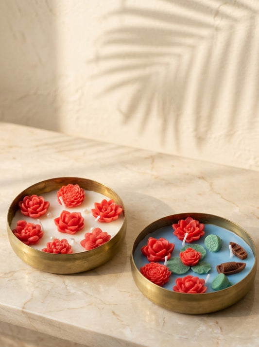 Two round brass candle holders with floral wax designs, one with red rose-shaped wax flowers on white wax and the other with red rose-shaped wax flowers, green leaves, and small brown boat-shaped wax accents on a light blue wax base, displayed on a beige marble surface.