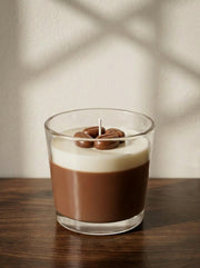 A candle in a clear glass jar sits on a dark wooden table against a light wall with soft window shadow patterns. The candle has a layered design resembling a coffee drink, with a rich brown lower section and a creamy off-white upper section, topped with three chocolate-coated coffee beans and a small white wick.