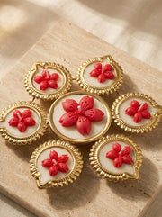 A set of six decorative brooches arranged in a flower-like cluster on a beige stone slab. Each brooch has a gold-toned scalloped frame, a white oval center, and a raised red flower motif.