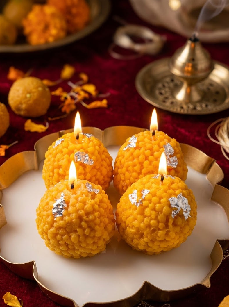 Four lit yellow marigold candles decorated with silver foil, arranged on a white and gold scalloped plate over a red fabric surface with traditional decorative elements.