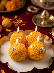 Four lit yellow marigold candles decorated with silver foil, arranged on a white and gold scalloped plate over a red fabric surface with traditional decorative elements.