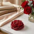 Red rose-shaped candle on a marble surface with books and a vase of red roses in the background.