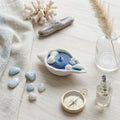 Decorative setting with a shell-shaped candle, stones, a compass, and a bottle of perfume on a light surface.