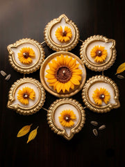 A decorative arrangement of gold-toned diya-style holders on a dark wooden surface, each containing a yellow flower, with one larger flower in the center and several scattered seed-like accents around.