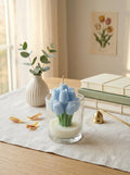 Candle shaped like a cactus in a glass container on a table with books and a vase in the background.