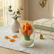Candle shaped like orange tulips in a glass container on a table with books and a vase in the background.