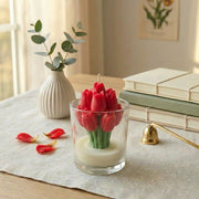 Red tulip-shaped candle in a glass container on a table with books and a vase in the background.